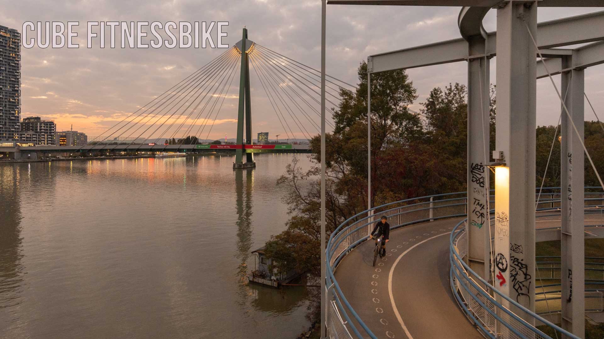 The image shows a city by a river. In the foreground, a cyclist is riding along the bike path at dusk. The image is captioned