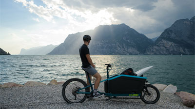 A man is standing with his cargo bike on the lakeshore. He is looking at the mountains in the background.