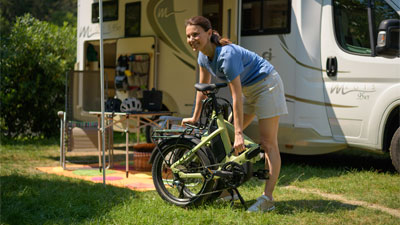 A woman is standing in front of a motorhome, in front of her is a folded green folding bike.