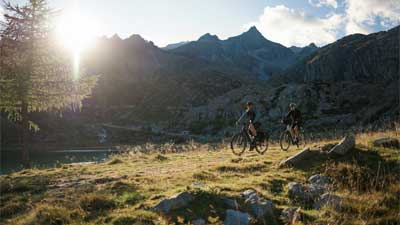 A couple rides their trekking bikes on a hiking trail. In the background are mountains with the sun peeking out from behind them.