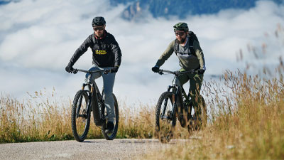 Two cyclists are riding their e-mountain bikes on a bike path. Mountains can be seen in the background.