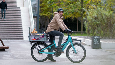 A cyclist rides through the city center on his blue Cube Longtail. He has a box of groceries stored on the luggage rack.