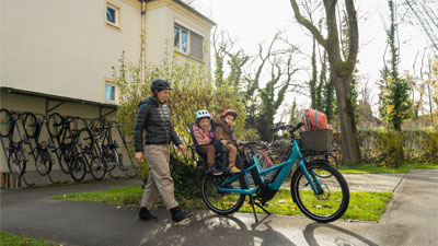 A mother is seen walking toward her blue cargo bike. Two small children are sitting on the luggage rack. The scene is set in a residential area.