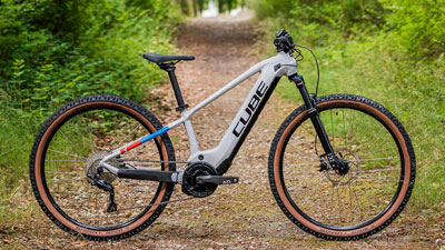The image shows a white Cube children's e-bike with small blue and red details. It is standing on an overgrown forest path.
