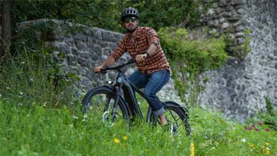 A cyclist rides his mountain bike across a meadow, with a stone wall in the background.