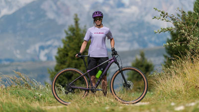 A woman in purple cycling gear is standing in front of a purple Cube mountain bike. A mountainous landscape can be seen in the background.