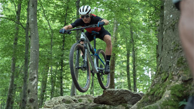 A cyclist is riding through the forest and jumps off a rock.