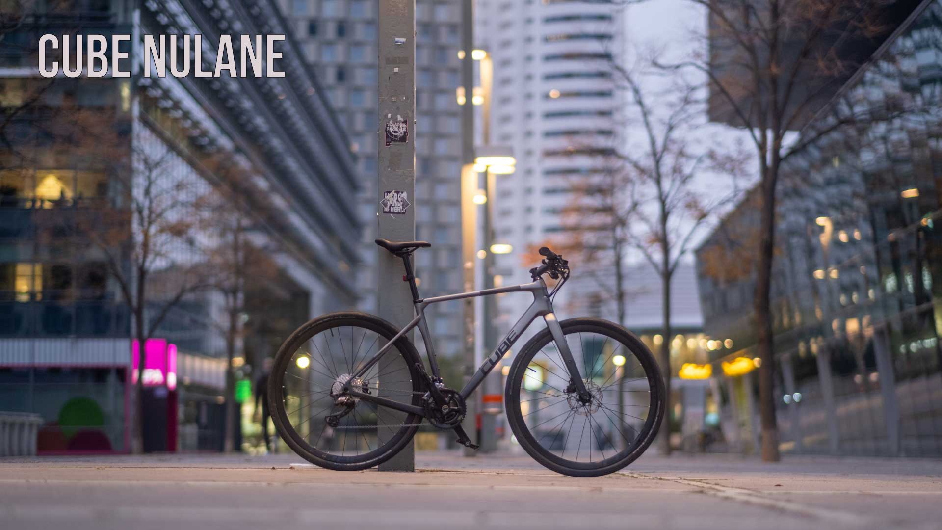 Banner with the words Cube Nulane. A Cube Nulane exercise bike is leaning against a pillar in the city center.