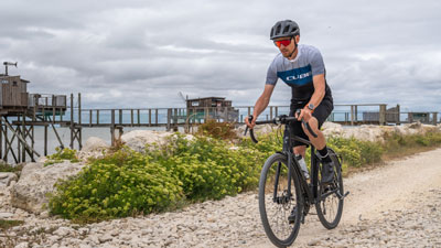 A man in cycling gear can be seen riding his gravel bike on a beach path.