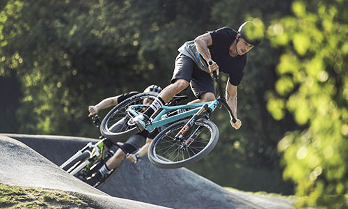 Cyclist jumps with a turquoise dirt bike in a bike park, another cyclist with a green bike can be seen in the background.