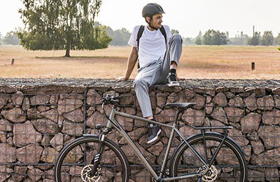 A man wearing a helmet is sitting on a wall. In the foreground is a gray bicycle, and in the background is a field landscape.
