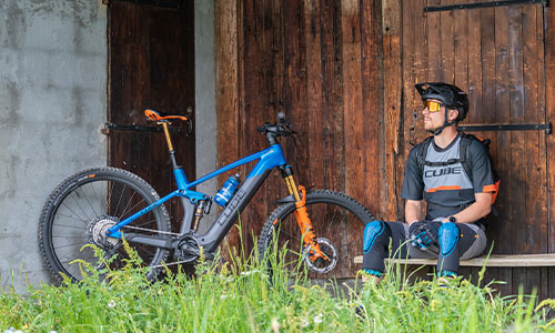 A cyclist is sitting on a bench in front of a barn. Next to him is his Fully e-mountain bike.