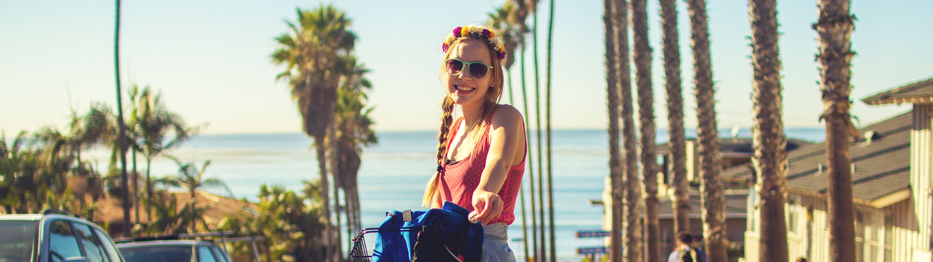 Smiling woman standing next to a bicycle, holding the handlebars, with palm trees and the sea in the background.