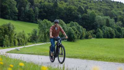 A cyclist rides his mountain bike along a paved bike path with a rural landscape in the background.