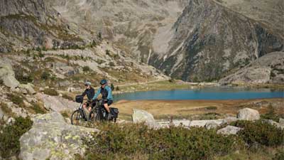 A couple rides trekking bikes through rocky terrain, with mountains and a lake visible in the background.