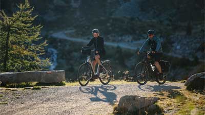 A couple rides mountain bikes on a bike path with a mountainous landscape in the background.