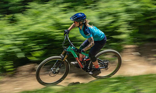 You can see a woman on a mountain bike riding quickly along a forest path.
