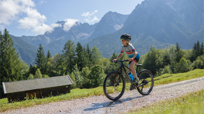 A boy rides his mountain bike on a gravel path through a mountainous landscape. A hut can be seen in the background.