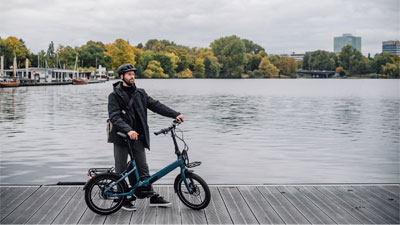 A man is standing next to his blue folding bike on a jetty. In the background, you can see water and a city.