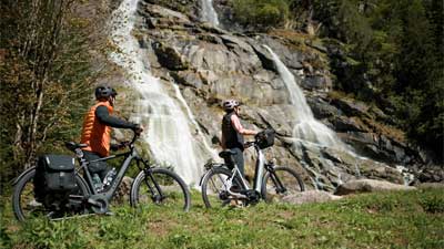 A couple pushes their bicycles across a meadow, with a waterfall in the background.