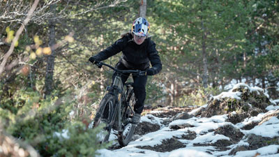 A cyclist rides her mountain bike through the uneven forest, where the ground is still partially covered with snow.