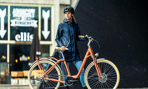 A woman stands behind her orange city bike in the city center.